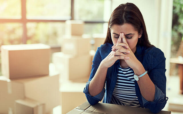 A young woman has her hands frustratedly clasped together in front of her face with her elbows resting on a moving box with additional packed moving boxes sitting on the floor in her home's living room behind her with the sun shining through the windows