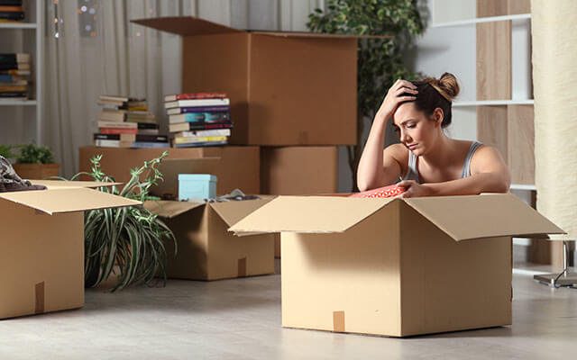 A woman sits on the floor of her home with an open moving box in front of her looking defeated, as more moving boxes and belongings sit in the background