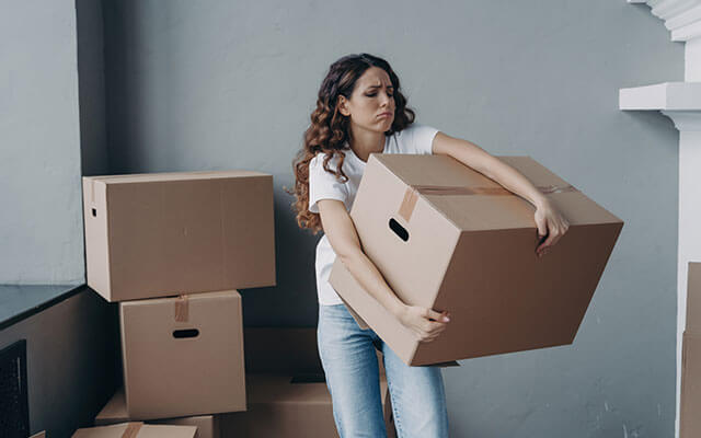 A woman struggles to hold onto a large, taped up moving box inside of her home