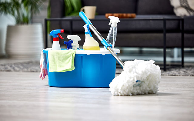 A view of a number of cleaning supplies in a cleaning bucket with a duster sitting on the bucket on the floor of a nice suburban home