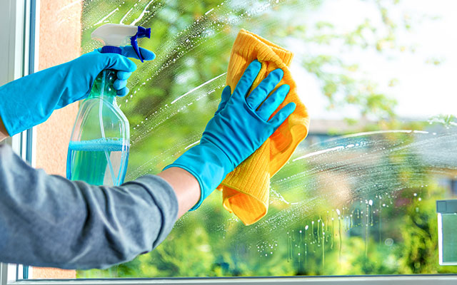 A person wearing cleaning gloves uses a microfiber cloth and a glass cleaning solution cleans a window in their home