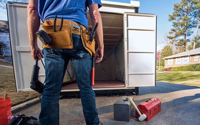 A man with a tool belt holding a power drill surrounded by tool boxes and additional tools stands on a neighborhood street in front of an open 1-800-PACK-RAT portable storage container ready to get to work on an under construction home