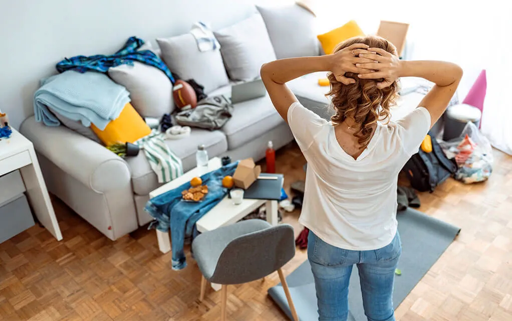 A woman faces her living room's couch that has a bunch of clothes and toys strewn about it, and on the floor