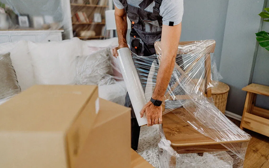 A laborer helping move someone's belongings wraps a chair in some protective wrapping paper for the journey in a living room