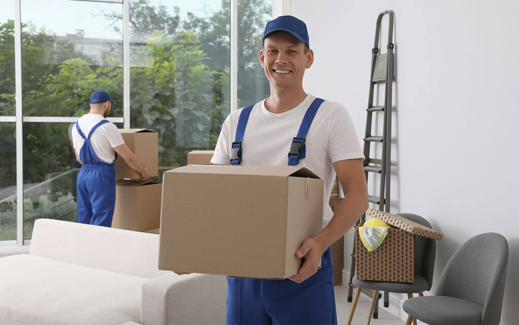 A professional mover laborer holds a box in his arms as he colleague in the background grabs another box to carry out of the house