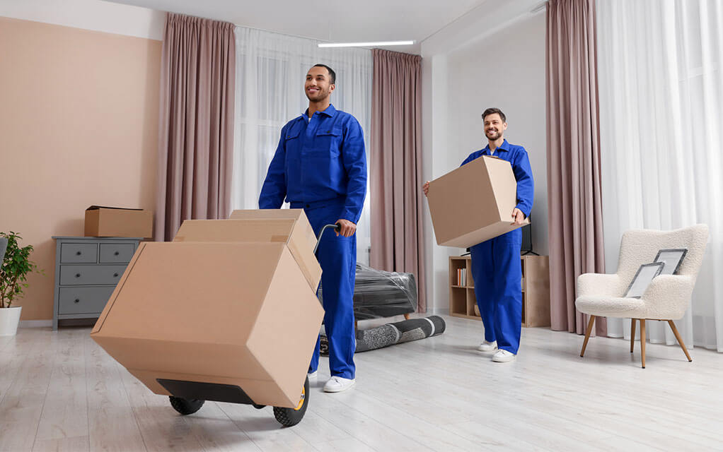 Two professional laborers, one pushing a hand cart with large boxes on it and the other carrying a box, move items out of a customer's home