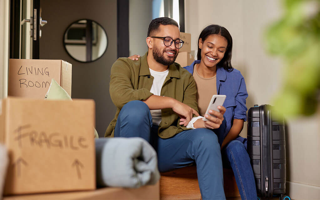 A couple sits on their home's floor and look at some on the man's phone as labeled moving boxes sit in both the background and the foreground