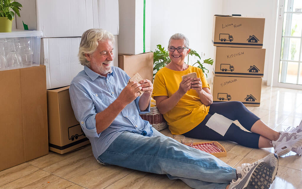 An older couple sits on the floor of their home eating some sandwiches and are surrounded by packed boxes that are labeled for their move