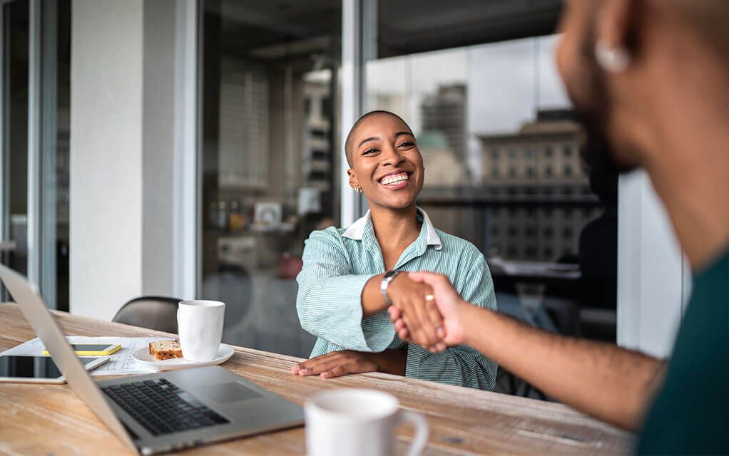 A young woman sits at a conference table with a laptop open in front of her and a white coffee cup on a plate, shaking hands with a man sitting to her left, smiling as she does so