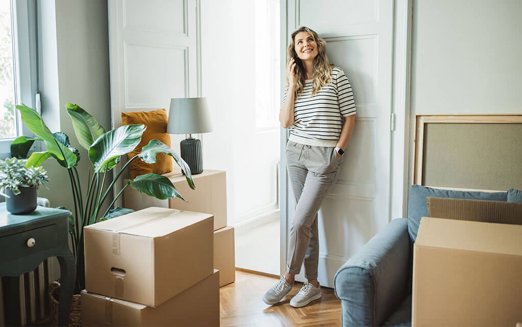 A young woman stands against the wall of her living room and is surrounded by packed boxes and other items ready to be loaded for a move