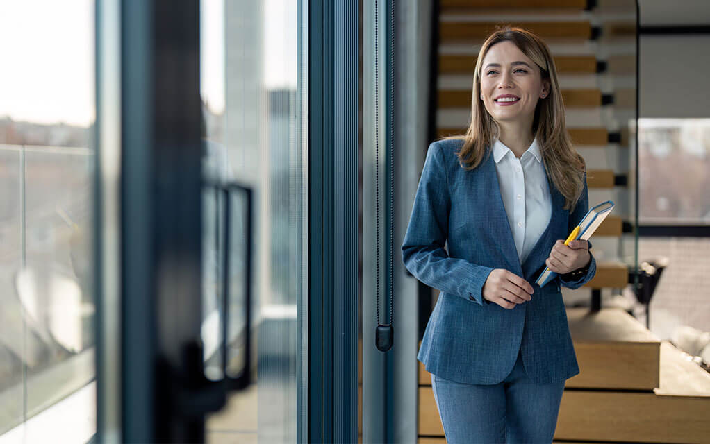 A young professional woman wearing a blue suit smiles as she holds some notebooks and looks on through the office windows