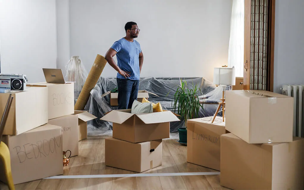 A young man stands with his hands on his hips, seemingly overwhelmed by the moving boxes and and chaos surrounding him as he tries to get his move going