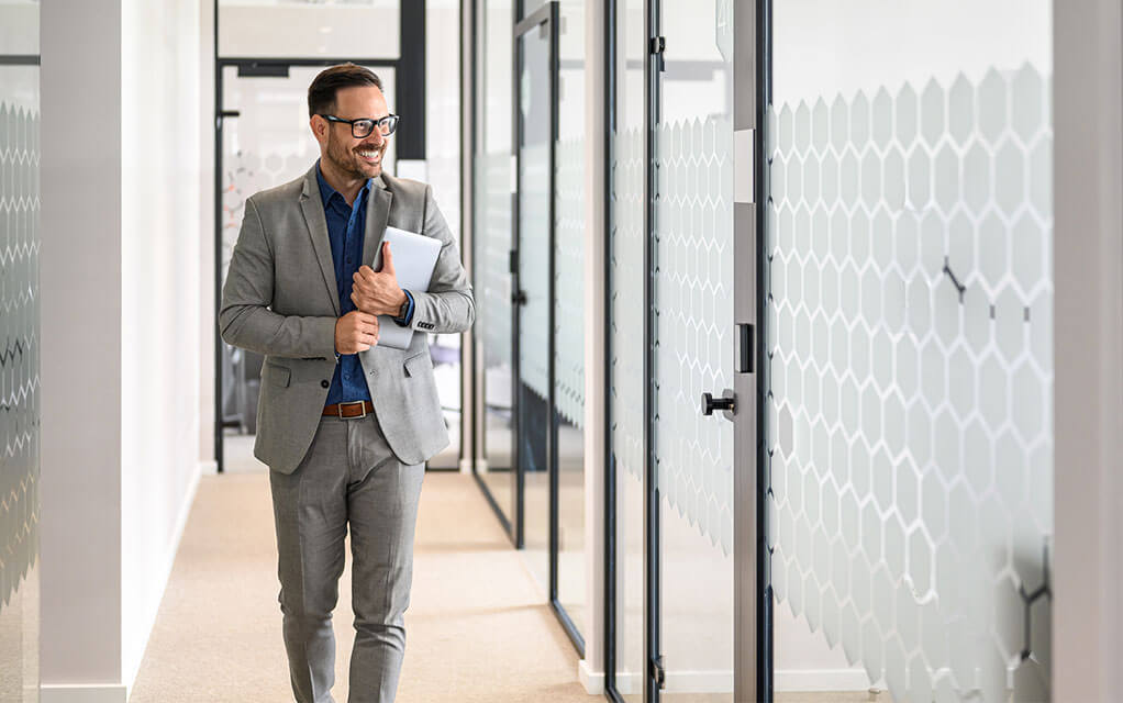 A man carrying some papers smiles as he walks down a hallway in his office as he looks to enter a conference space for a meeting