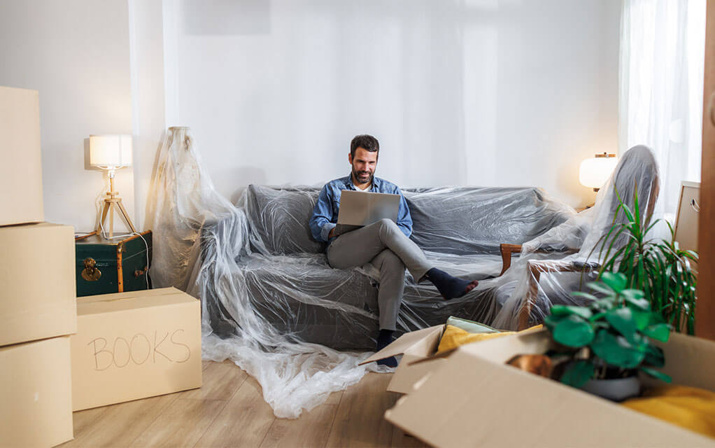 A man sits on his covered couch as he does work on his laptop computer, surrounded by packed and opened moving boxes in his living room