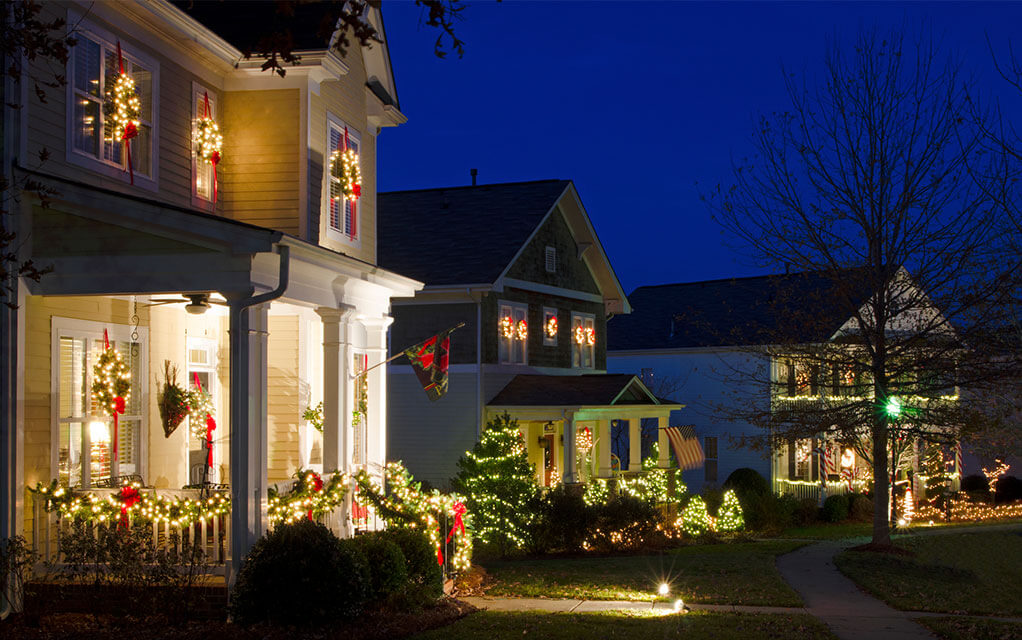 Several houses adorned in holiday and Christmas decorations and lights sit on a quiet suburban neighborhood street on a winter night