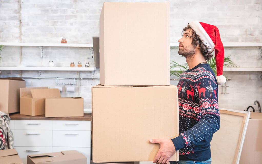 A man wearing a Santa hat holds two large boxes stacked on top of each other in his home's kitchen surrounded by additional moving boxes and items