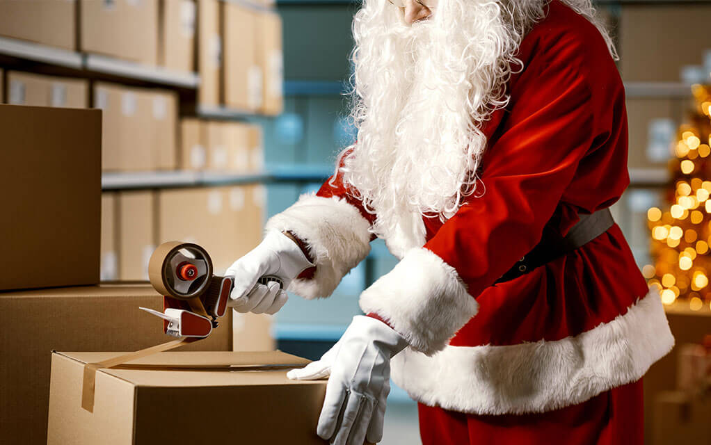 Santa in his iconic red outfit closes a moving box with some tape from a tape gun, as additional boxes sit in the background waiting to be moved
