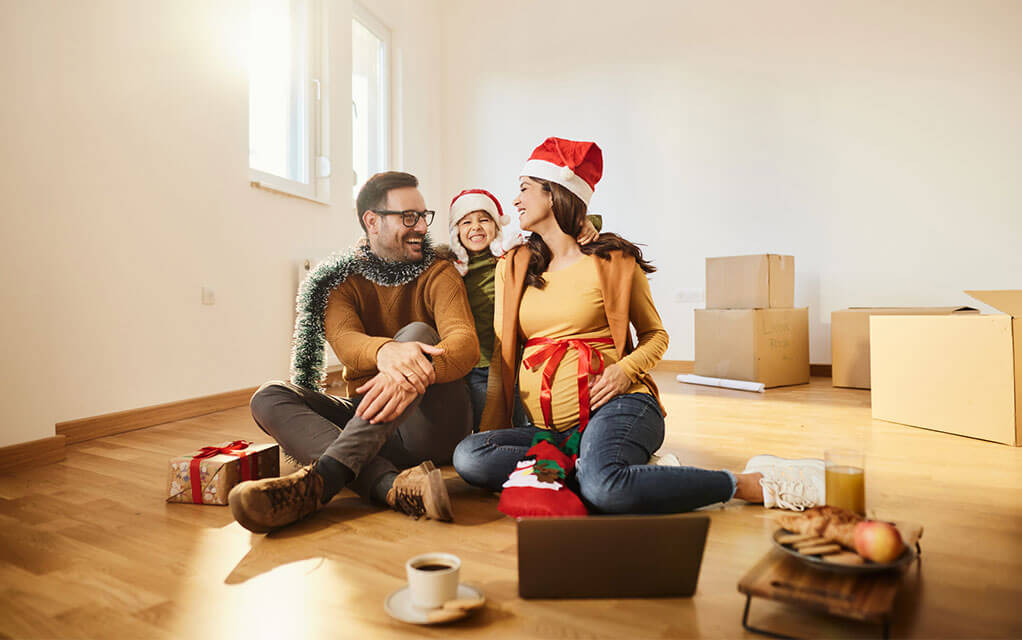 A happy man sits on the floor with his pregnant wife as their young daughter stands behind them as an open laptop and coffee cup sit in front of them with moving boxes sitting in the background