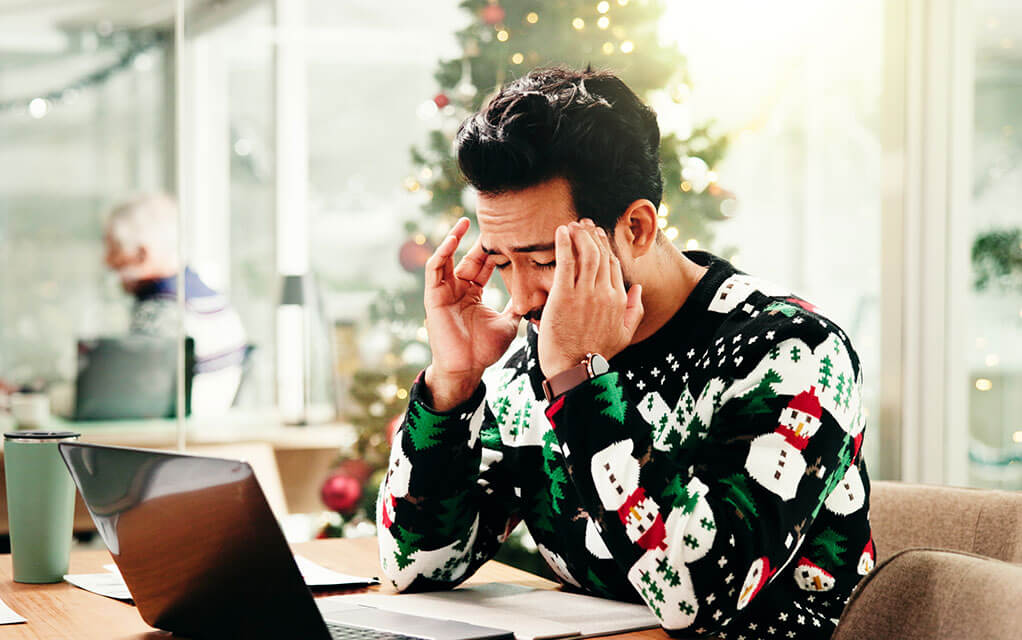A stressed out man wearing a Christmas sweater sits in front of his laptop computer rubbing his temples