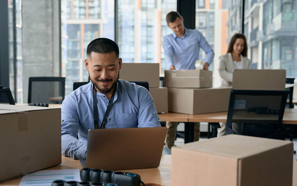 A man sits at an office table doing work on his laptop with moving boxes surrounding him with two more colleagues in the background packing boxes for an office move
