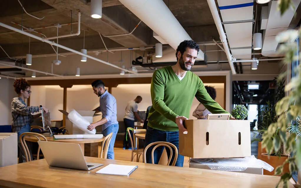A young man places a box down on a table in a new office space, as he and his fellow employees begin to arrange their new office