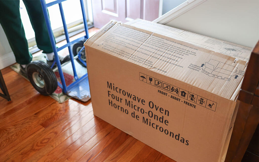 View of a rectangular cardboard box on the floor of a home with "Microwave Oven" on it as a person with a moving dolly is getting ready to move it out of the house