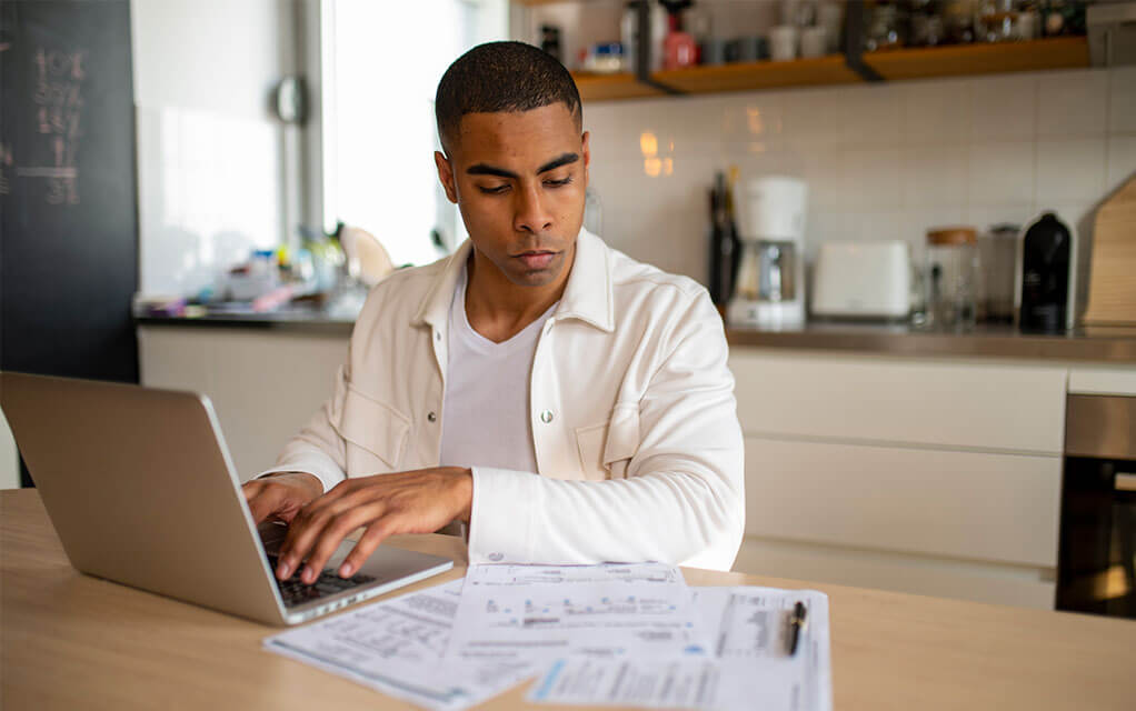 A young man sits at his kitchen table in his kitchen, doing work on his laptop as he looks at some important documents set out on the table