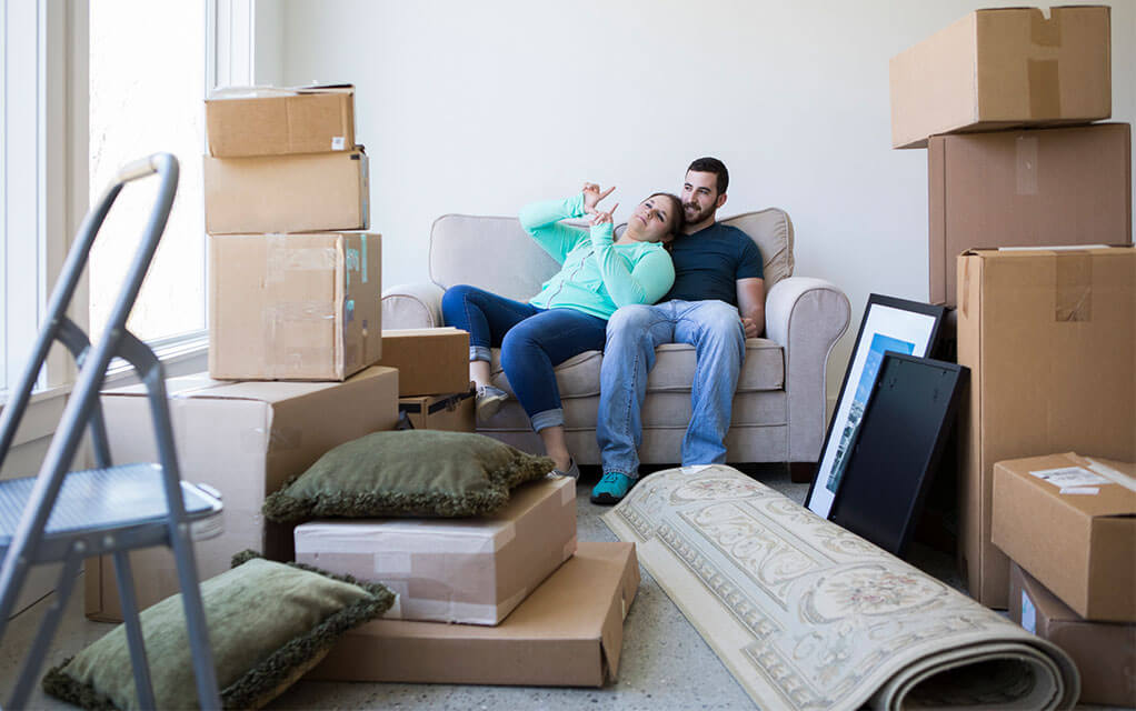 A happy couple sits on their couch, as moving boxes stacked on top of each other and other belongings and furniture sit around them waiting to be moved in their living room
