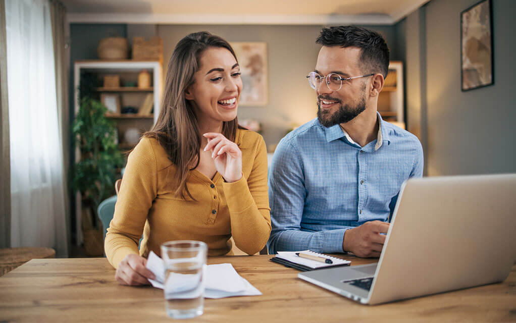 A young couple sitting at their kitchen table jovially talk to each other as they do research for a project on their laptop as the woman takes notes on some paper sitting on the table in front of her