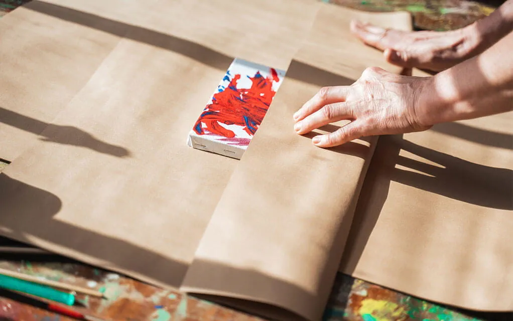 A person wraps a small canvas painting in some acidic free brown paper wrap to prepare for shipping