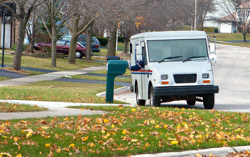 A US Mail truck is seen on a neighborhood street on a cloudy fall day as a mail carrier puts mail into a mail box