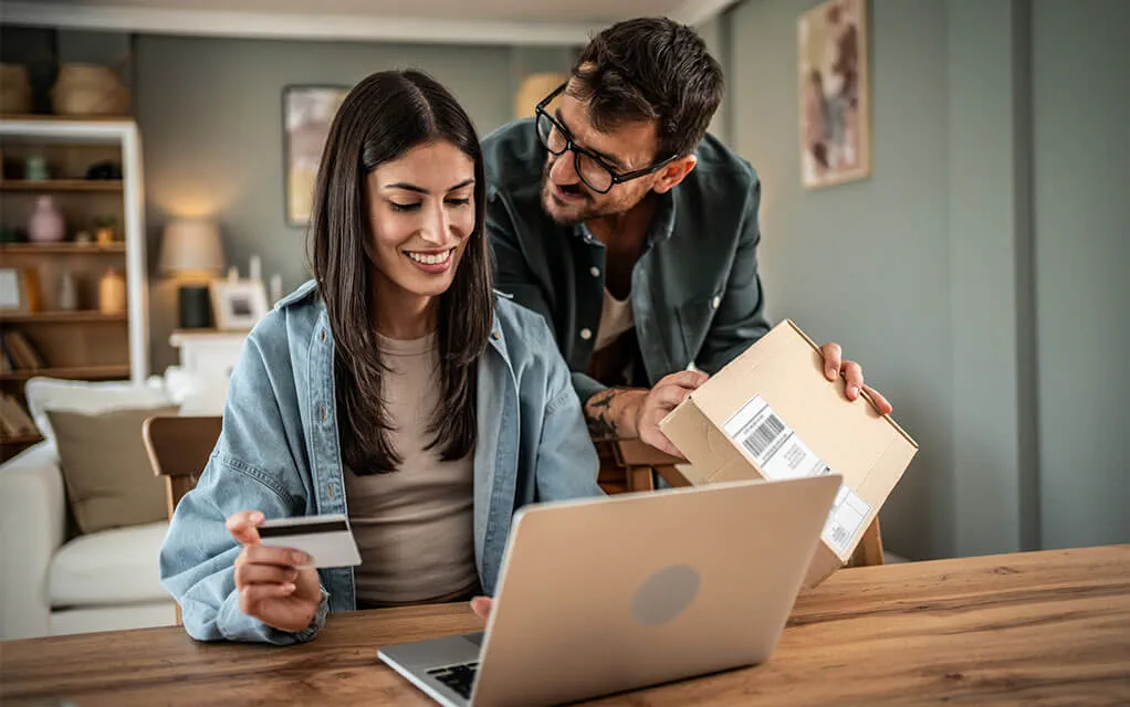 A couple makes a purchase on their laptop as the woman holds a credit card and inputs info on her laptop while her husband shows her a package that arrived