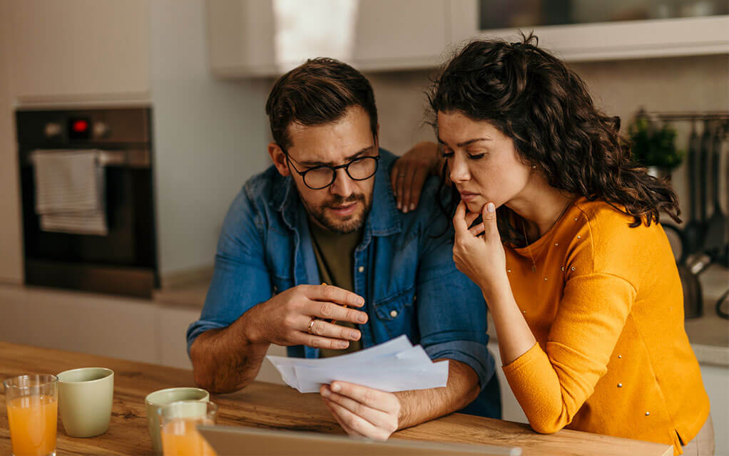A young couple looks at some mail with concerned faces as they stand at their kitchen island with two glasses of orange juice in front of them