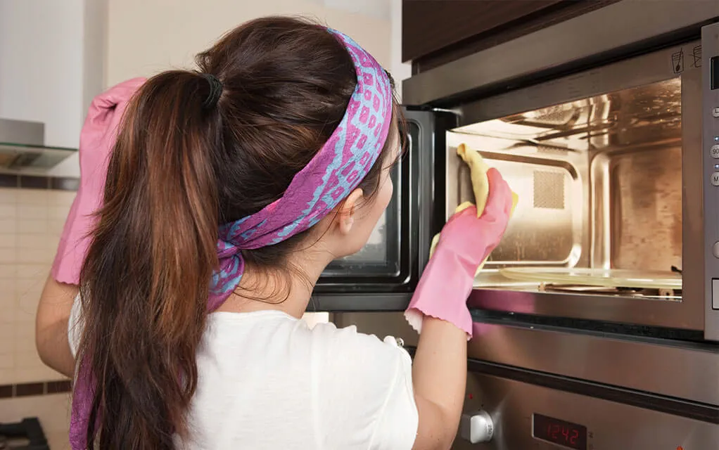 A woman cleans the inside of her home's microwave, using a microfiber cloth to scrub the inside while wearing pink gloves
