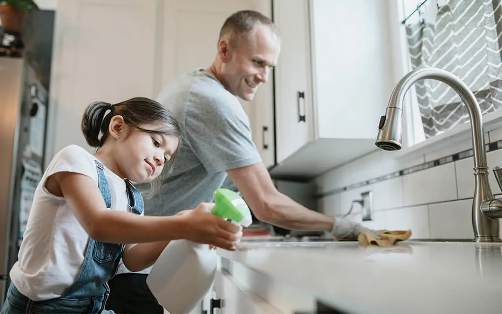 A father and his young daughter help clean their kitchen's counters as the daughter sprays a cleaning solution