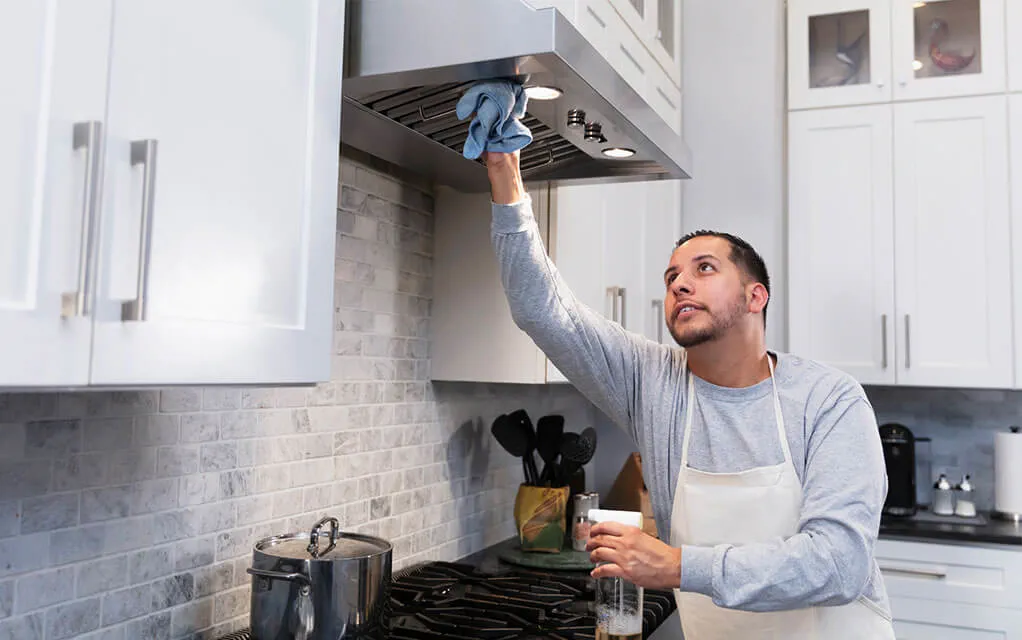 A man uses a cloth to clean the hood of his kitchen's stove, as he holds a bottle of cleaner in his other hand in the kitchen