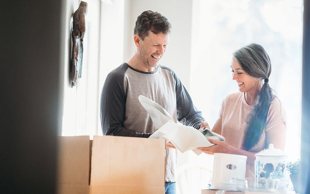 An older couple laughs together as they unpack items from boxes after moving into their new home on a bright, sunny day