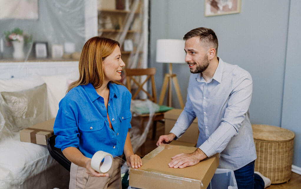 A young couple is packing boxes for an upcoming move, surrounded by other items and furniture needing packing