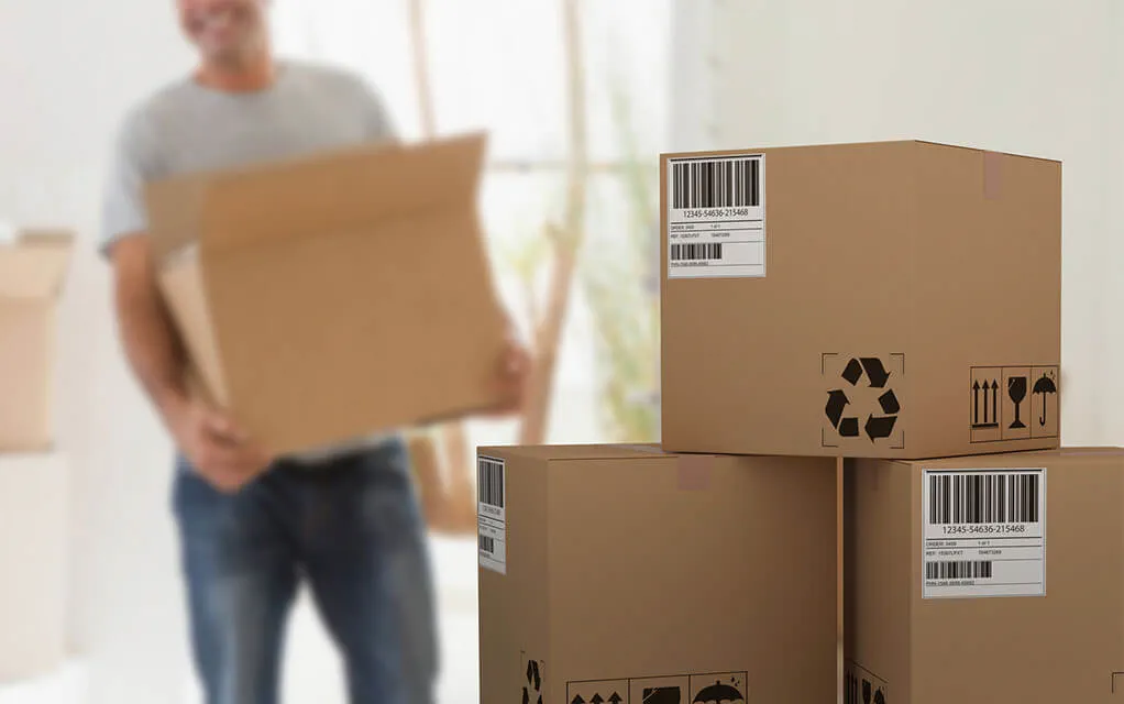 A stack of boxes sits on the floor, with one of the having the recycling symbol present on it as a man in the back ground carries a box