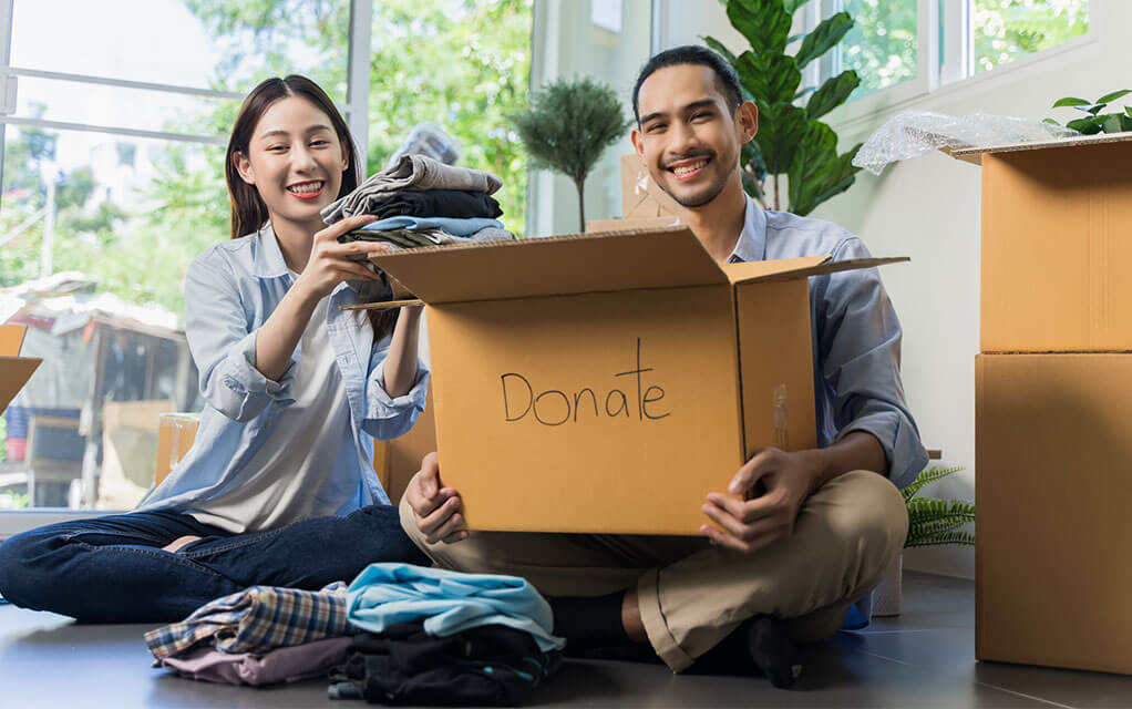 A couple smiles at the camera as the man holds a box with the word "Donate" written on it, while the woman places folded clothes into it