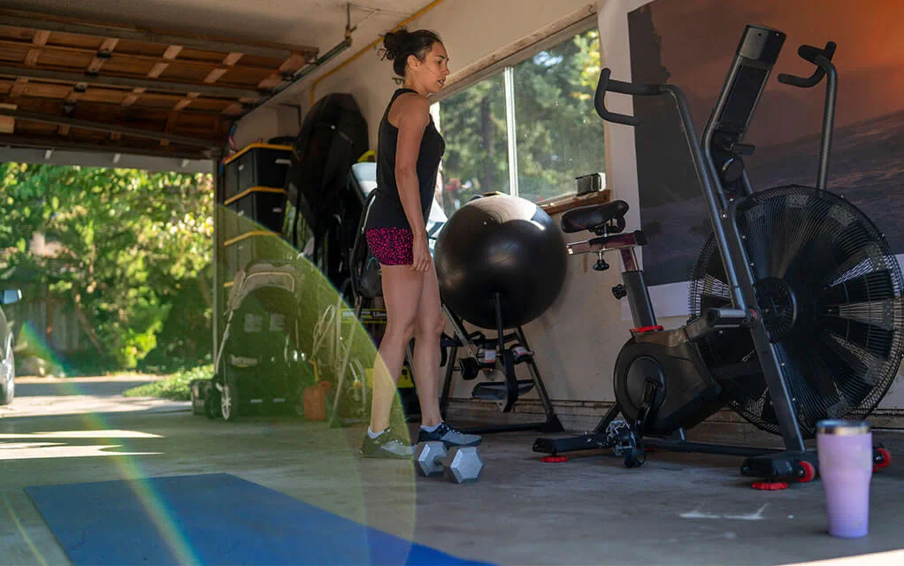 A woman stands in her garage with gym equipment and a medicine ball around her, with a dumbbell and a yoga mat on the floor