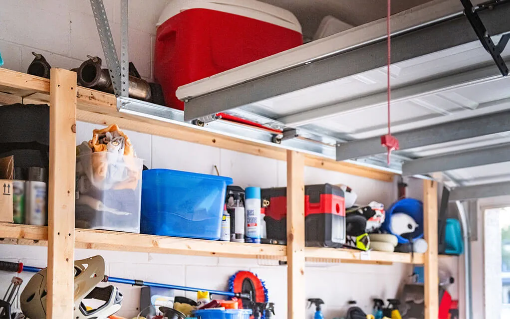 Wooden shelves along the wall of a garage hold bins, tools, and other household items