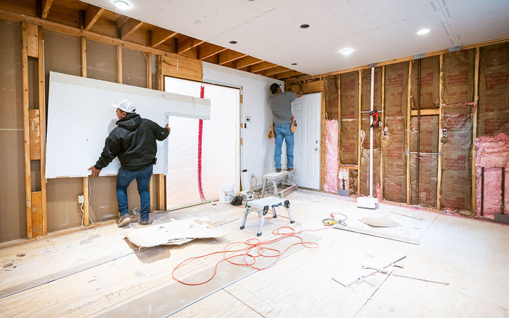 Two construction workers replace wall studs and drywall in a room undergoing a major renovation inside a home