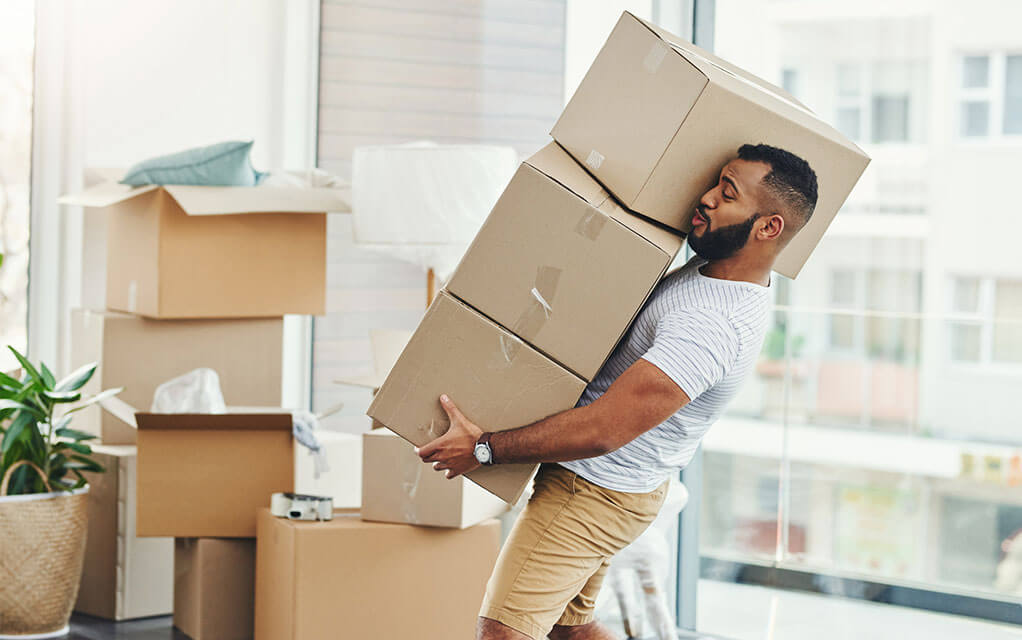 A man attempts to carry three packed cardboard boxes as the top box begins to slide off the stack and onto his shoulder