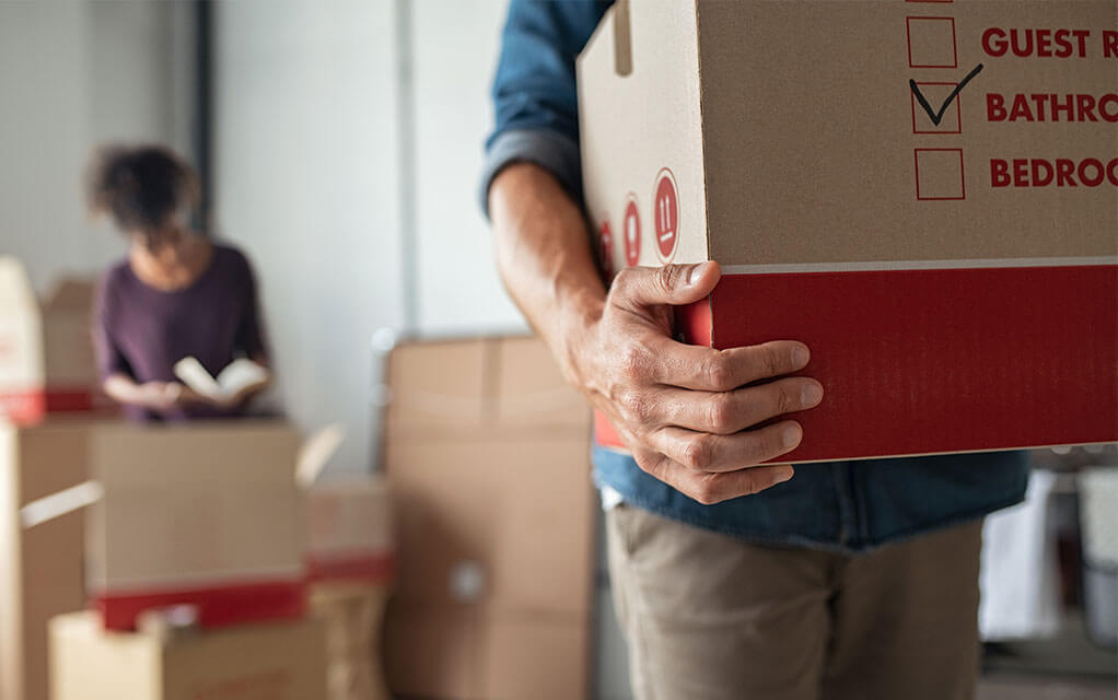 A man carries a box with a checkmark on a list of contents next to BATHROOM, as a woman in the background packs more moving boxes