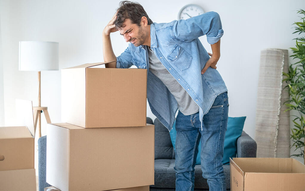 A man leans on a stack of moving boxes, squinting and holding his side, clearly in some sort of distress as he tries moving boxes in his living room