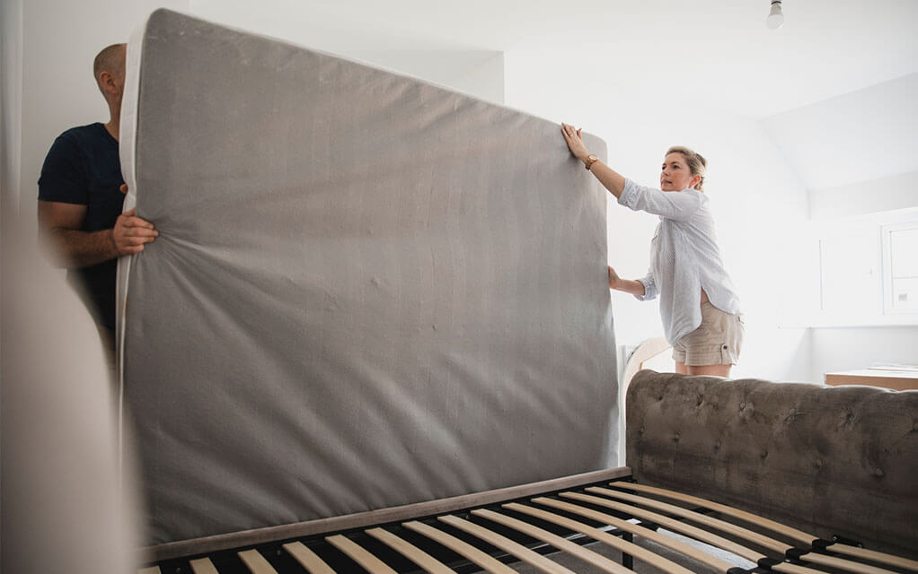 A man and a woman work together to get their mattress onto their bedframe in their room