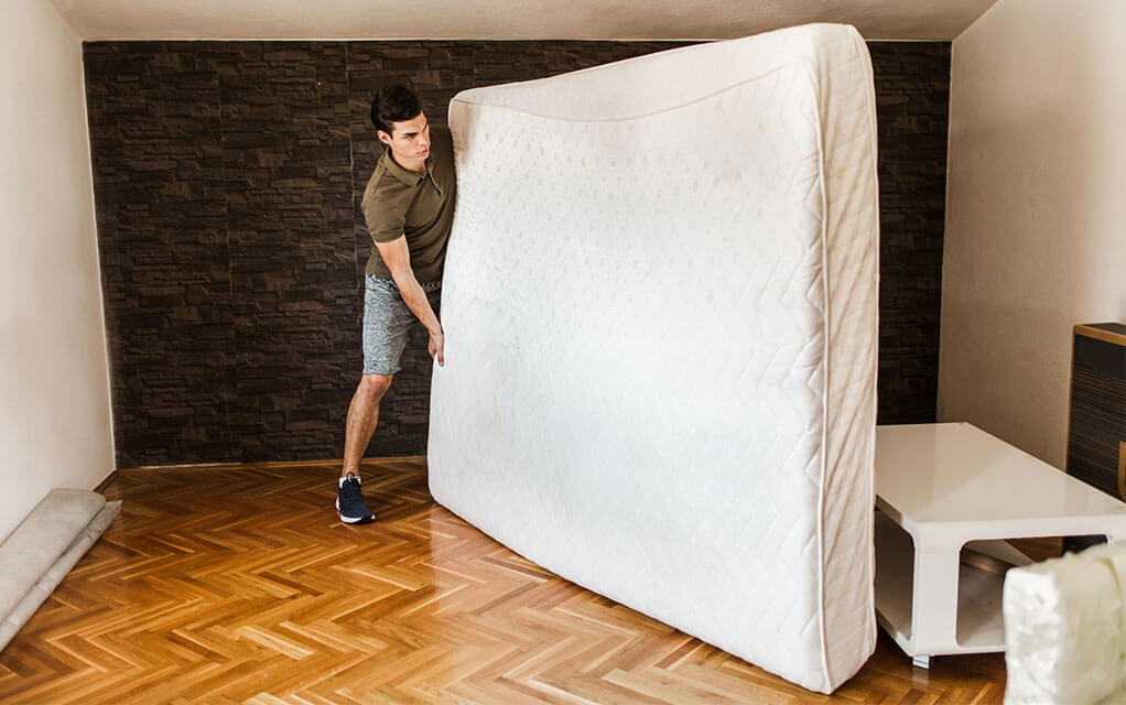 A man is moving a mattress out of his room on its side as it is covered in a mattress protector