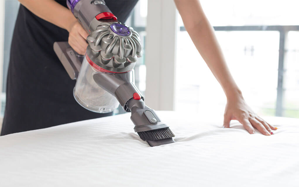 A person uses a battery powered fabric cleaning vacuum to clean the top of their mattress on a bright and sunny day