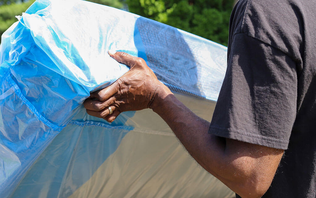 A man is holding up a mattress by the edge that is covered in a protective mattress cover outside as he moves it
